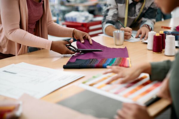 Close up of fashion designer cutting piece of textile while working at clothing workshop.